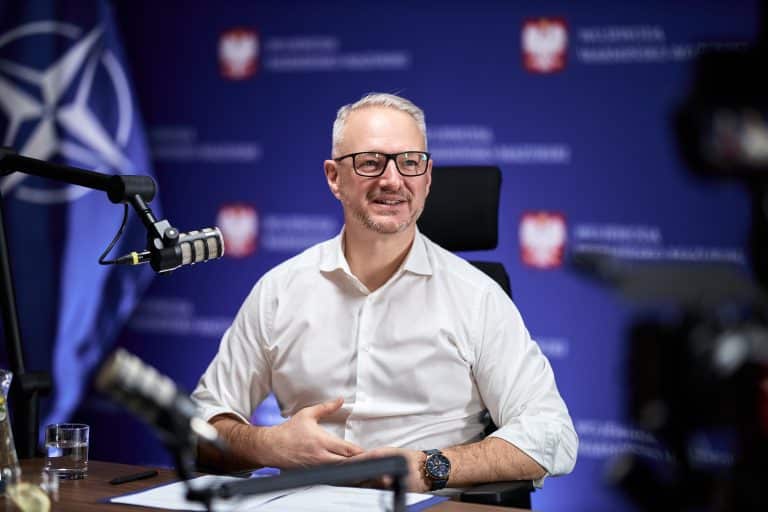 Wojewoda Radosław Król podczas konferencji, za nim logo województwa warmińsko-mazurskiego i symbol NATO.