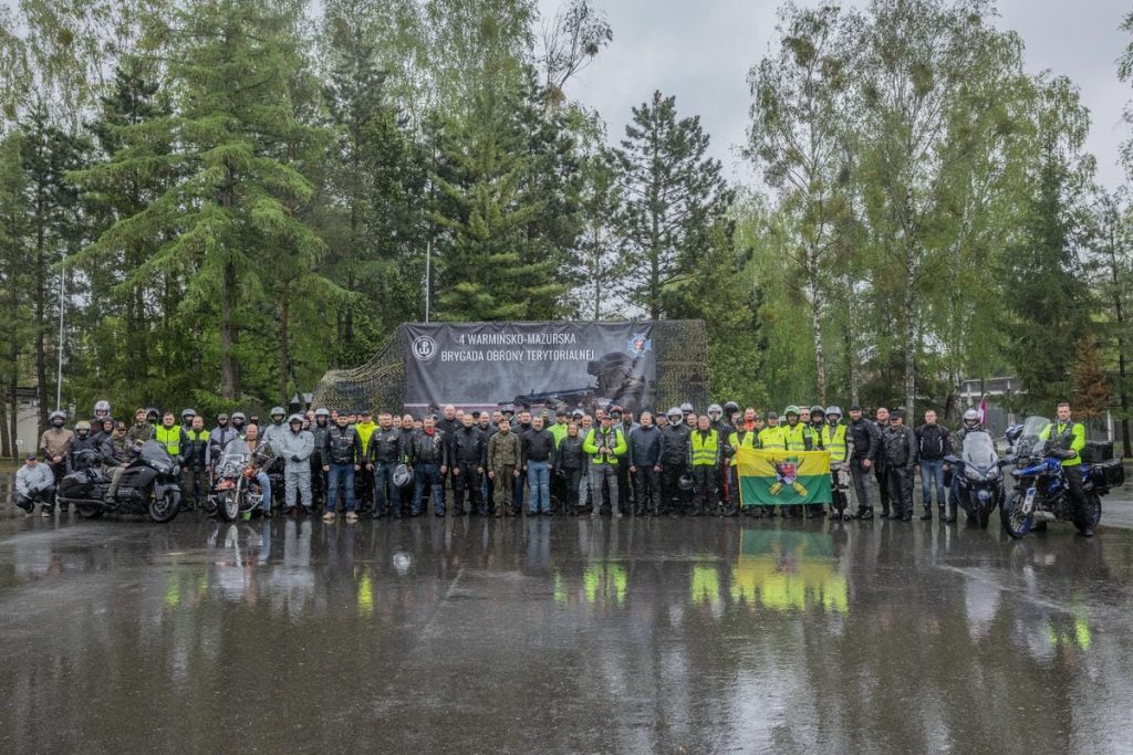 Grupa żołnierzy i motocyklist&oacute;w na uroczystości święta brygady w lesie, w deszczowej pogodzie.