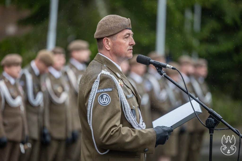 Żołnierz w mundurze podczas uroczystości święta brygady, z grupą żołnierzy w tle, na zewnątrz.