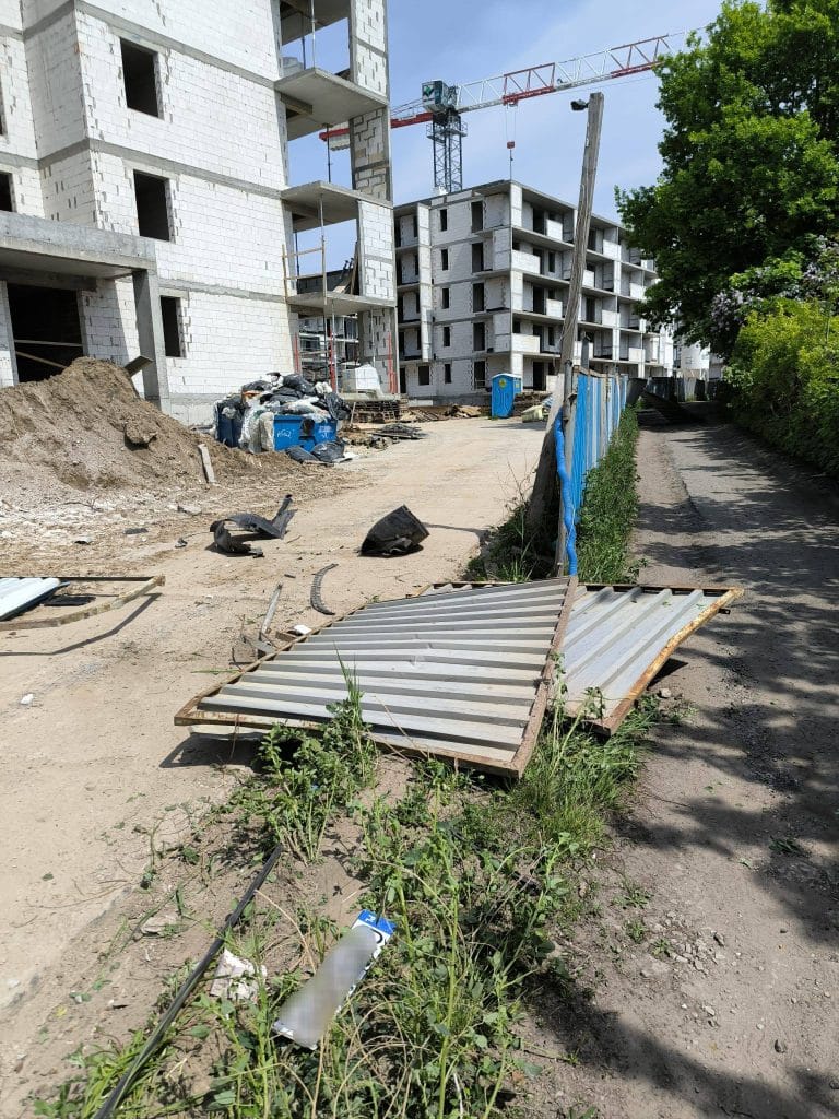 Zniszczone ogrodzenie placu budowy, fragmenty metalowych elementów i śmieci na drodze.