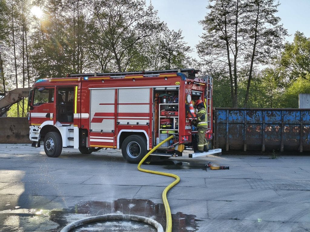Strażak gaszący pożar ciężar&oacute;wki na parkingu w Braniewie, w tle drzewa i dym.
