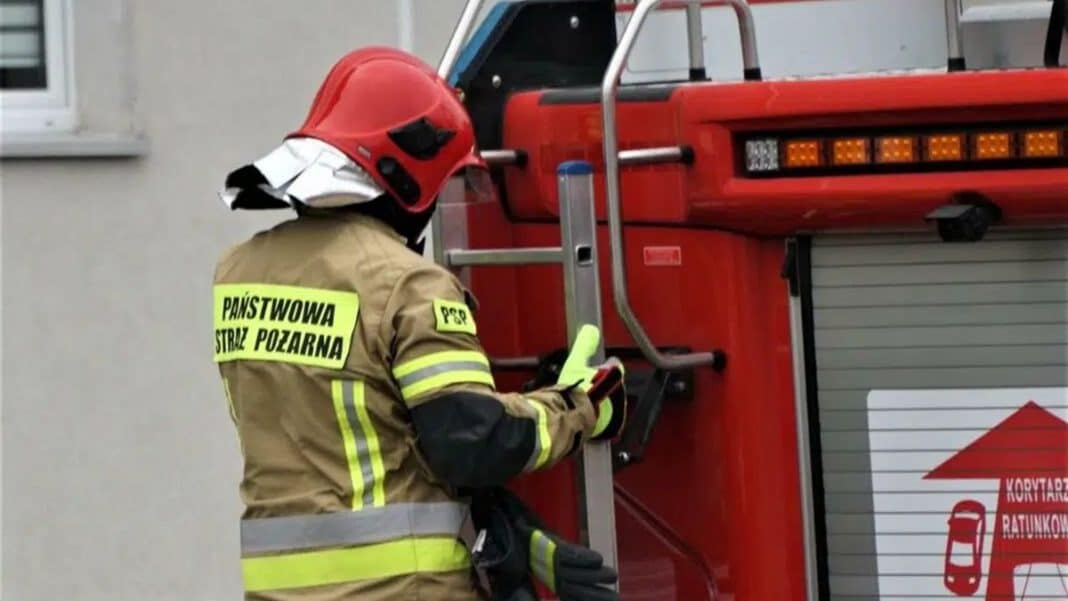 Strażak w hełmie i odblaskowej odzieży przy czerwonym wozie strażackim na miejscu zdarzenia.