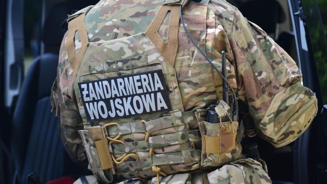 Żołnierz w kamuflażu z napisem Zandarmeria Wojskowa na plecach, stojący przy wojskowym pojeździe.