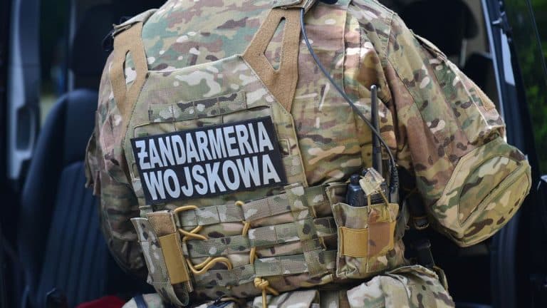 Żołnierz w kamuflażu z napisem Zandarmeria Wojskowa na plecach, stojący przy wojskowym pojeździe.