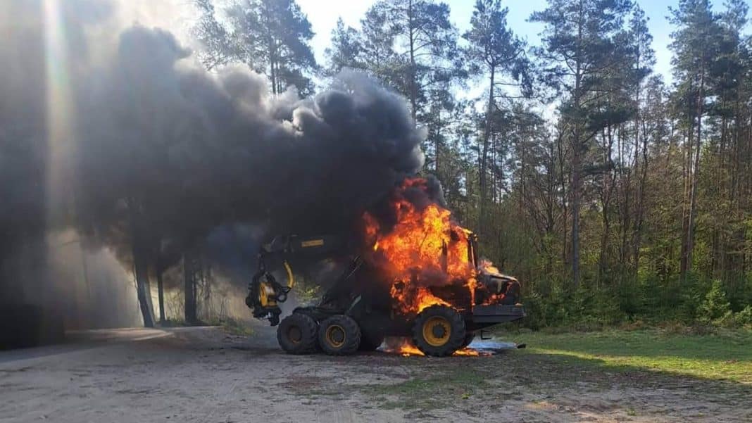 Spalony ciężki sprzęt leżący na drodze w lesie, z widocznym dymem i płomieniami.