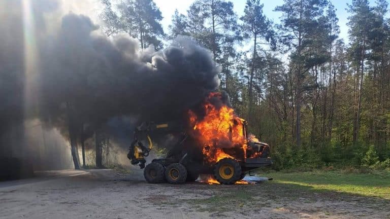 Spalony ciężki sprzęt leżący na drodze w lesie, z widocznym dymem i płomieniami.