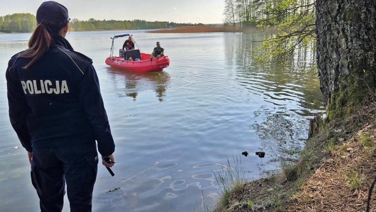 Policjantka nad jeziorem podczas akcji poszukiwawczej na Mazurach, w tle łódka z ratownikami.