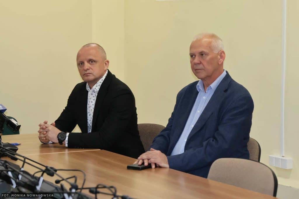 Dw&oacute;ch mężczyzn siedzi przy stole podczas konferencji, w tle jasna ściana.