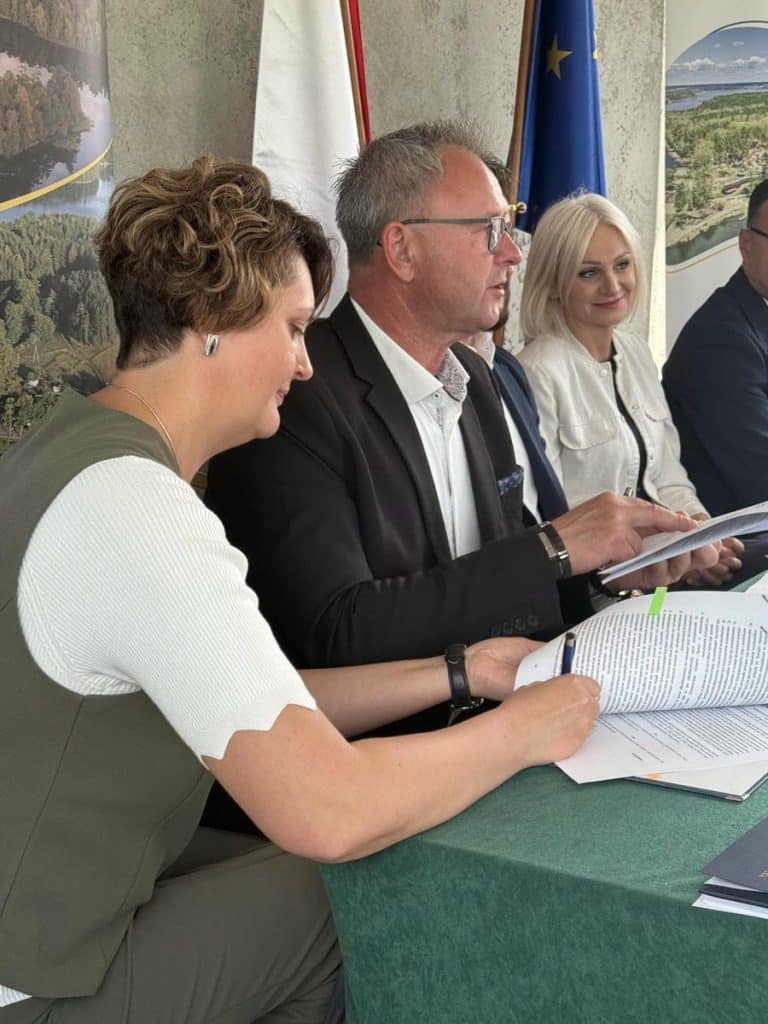 Uczestnicy spotkania przy stole, w tle flagi i widok na jezioro, podczas podpisania umowy na inwestycję.