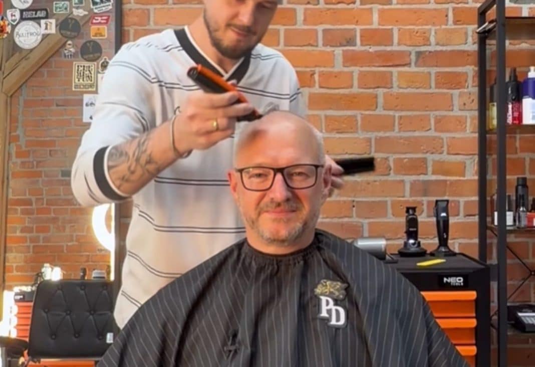 Wojewoda w salonie fryzjerskim podczas golenia głowy, za nim ceglana ściana i półki z kosmetykami.