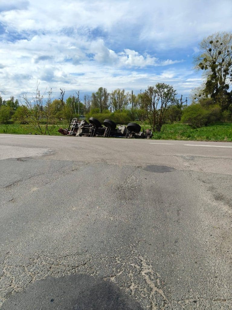 Przewr&oacute;cony dźwig leży na poboczu przy drodze, w tle drzewa i chmury na niebie.