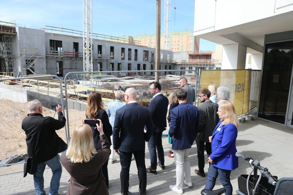 Grupa ludzi na zewnątrz, w tle budowa nowego szpitala, osoby robią zdjęcia i rozmawiają.