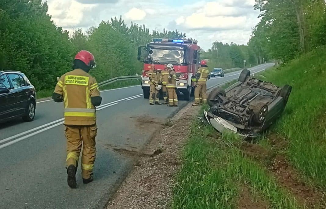 Rozbity samoch&oacute;d leży na boku przy drodze, w pobliżu strażacy i policja na miejscu zdarzenia.
