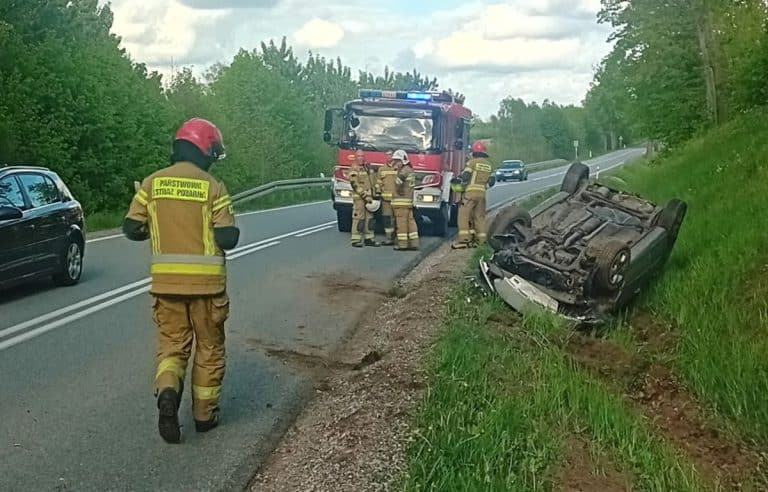 Rozbity samochód leży na boku przy drodze, w pobliżu strażacy i policja na miejscu zdarzenia.