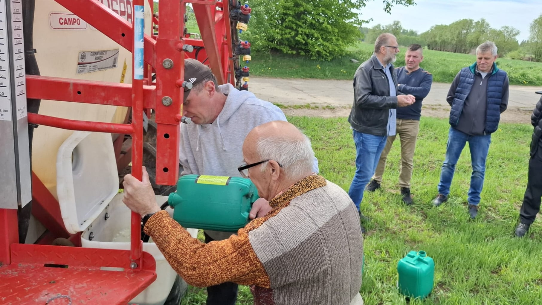 Grupa rolników podczas szkolenia na zewnątrz, z maszyną rolniczą i zbiornikami na wodę w tle.