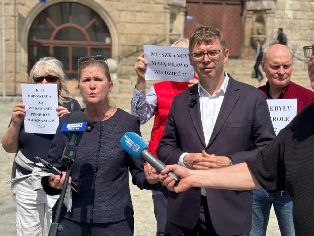 Grupa polityk&oacute;w na konferencji przed ratuszem, trzymająca plakaty i udzielająca wywiad&oacute;w.
