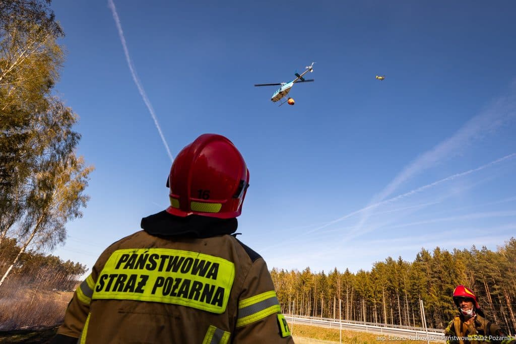 Strażacy nad lasem podczas akcji gaśniczej po pożarze na Mazurach, w tle helikopter i niebo.
