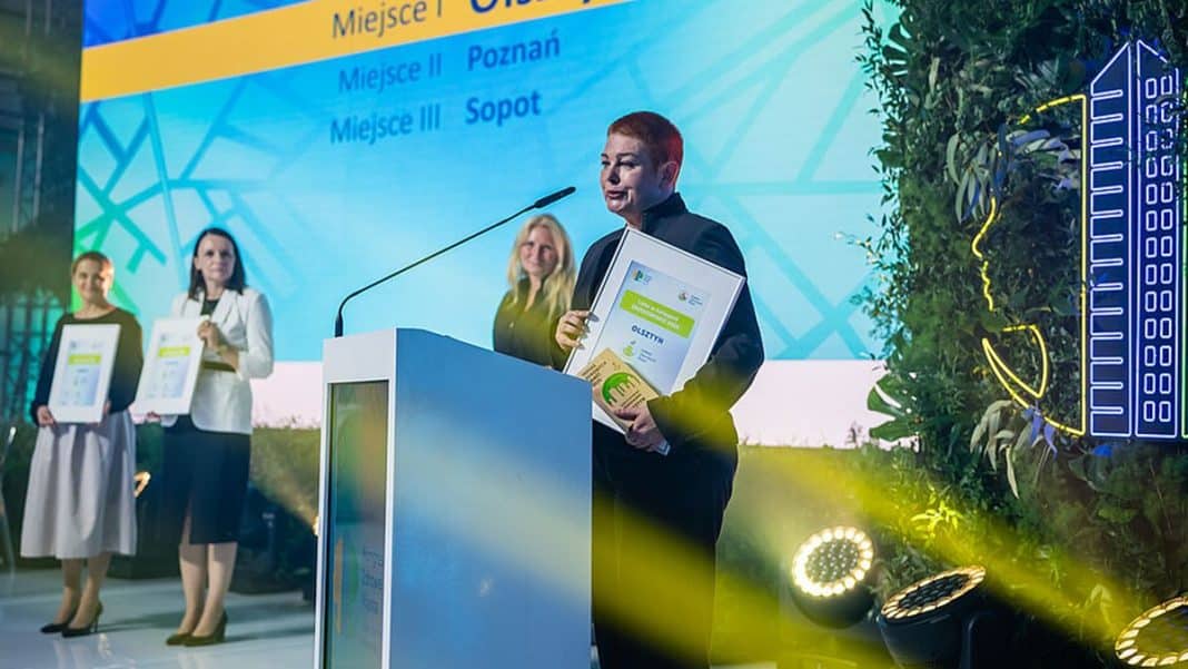 Osoba odbierająca nagrodę na scenie podczas ceremonii wręczenia, z dużym ekranem w tle.