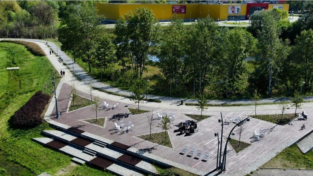 Widok na park z nowymi nasadzeniami drzew i miejscami do odpoczynku, w tle budynek z kolorową elewacją.