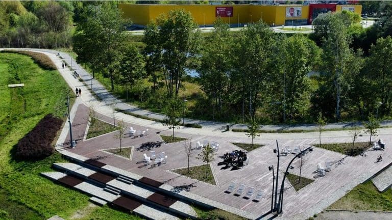 Widok na park z nowymi nasadzeniami drzew i miejscami do odpoczynku, w tle budynek z kolorową elewacją.