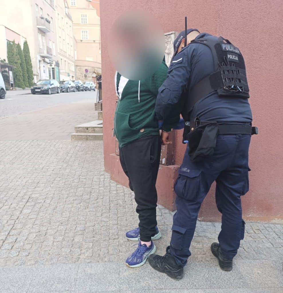 Policjant zatrzymuje mężczyznę na ulicy, oboje stoją przy ścianie budynku.