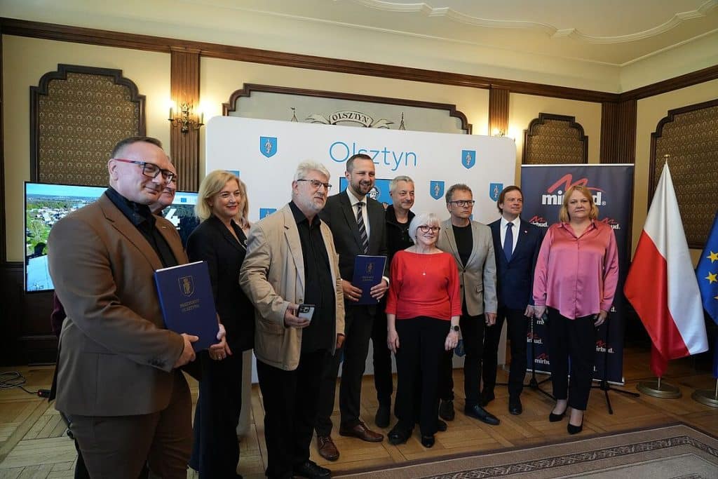 Grupa os&oacute;b podczas podpisania umowy na inwestycję drogową w Olsztynie, w tle logo miasta i flagi.