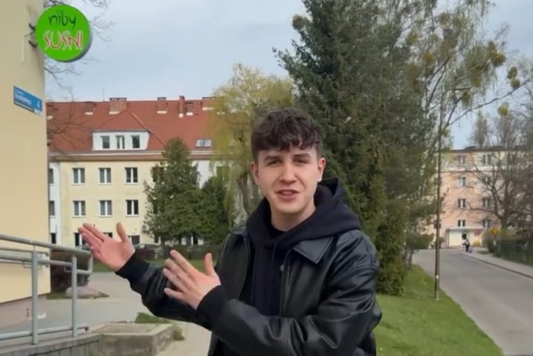 Młody mężczyzna na zewnątrz, gestykulujący rękami, w tle drzewa i budynki mieszkalne.