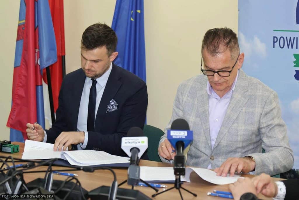 Dwa mężczyźni podczas podpisywania um&oacute;w na inwestycje drogowe, z flagami w tle.
