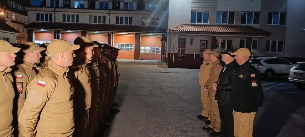 Grupa strażaków w uniformach stojąca w rzędzie na parkingu nocą, przygotowani do akcji gaśniczej.