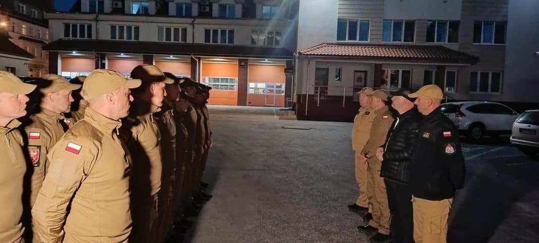 Grupa strażaków w uniformach stojąca w rzędzie na parkingu nocą, przygotowani do akcji gaśniczej.