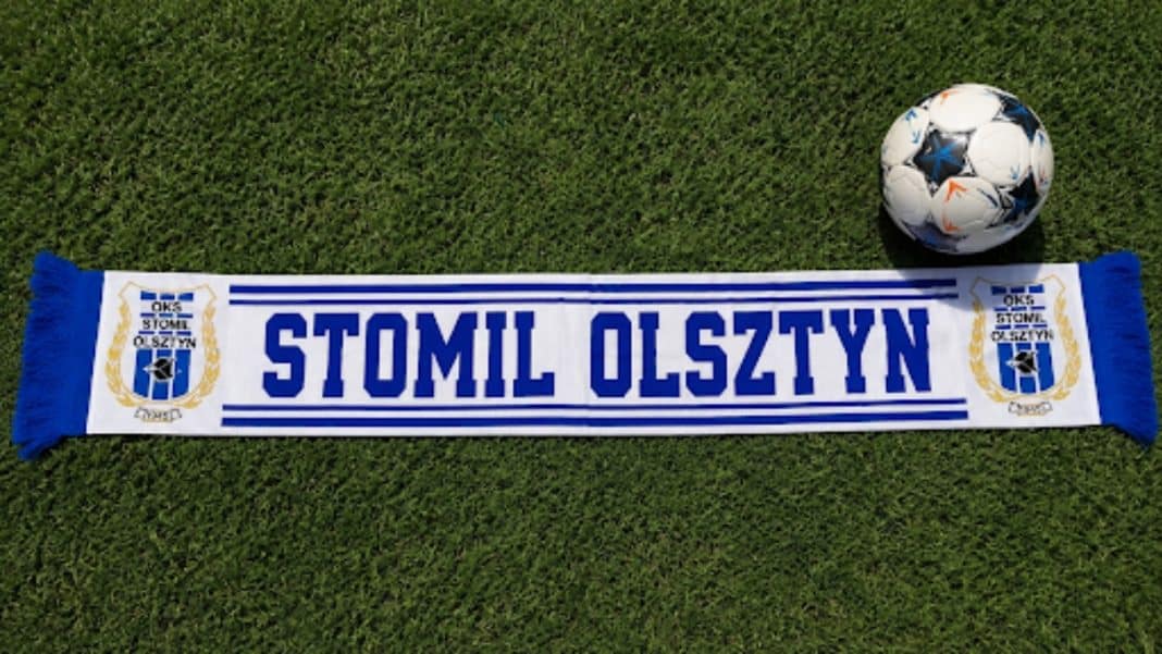 Szalik Stomilu Olsztyn i piłka ustawione na trawie, symbolizujące klub piłkarski.
