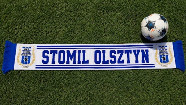 Szalik Stomilu Olsztyn i piłka ustawione na trawie, symbolizujące klub piłkarski.