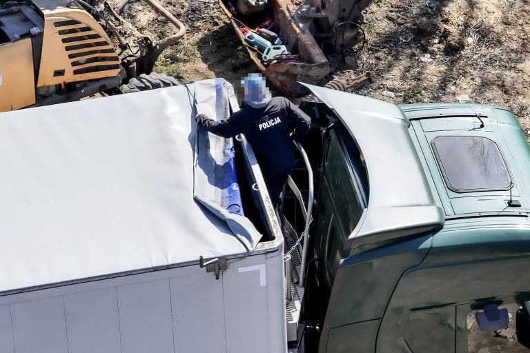 Policjant na dachu ciężar&oacute;wki podczas akcji na Mazurach, wok&oacute;ł zniszczone pojazdy i odpady.