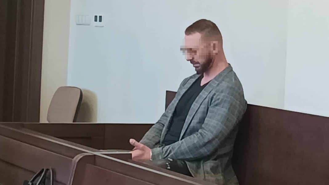 Mężczyzna w sali sądowej, siedzi na ławce, z głową pochyloną, z zakrytym wzrokiem twarzą.