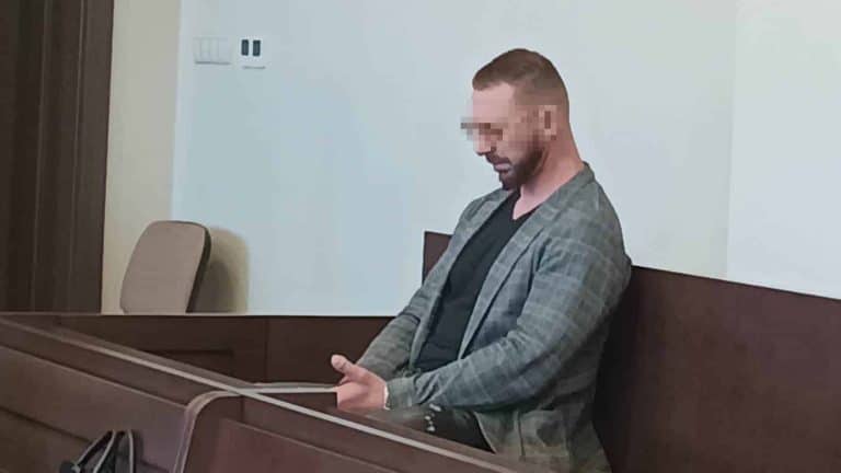 Mężczyzna w sali sądowej, siedzi na ławce, z głową pochyloną, z zakrytym wzrokiem twarzą.