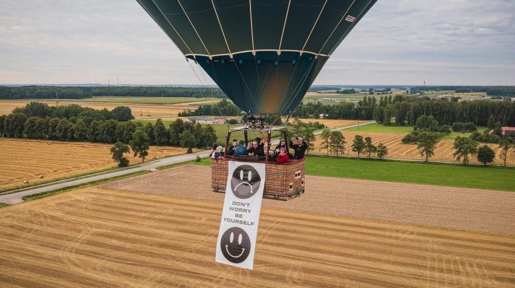 Balon na ogrzane powietrze z banerem z uśmiechniętym buźką i napisem "Don't worry be yourself" nad polami i lasami.