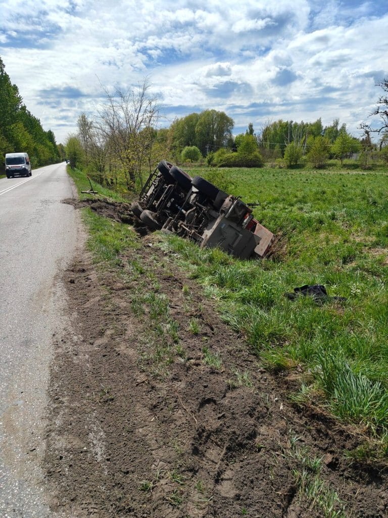 Przewr&oacute;cony dźwig leży na poboczu drogi wśr&oacute;d zieleni, w tle chmury na niebie.