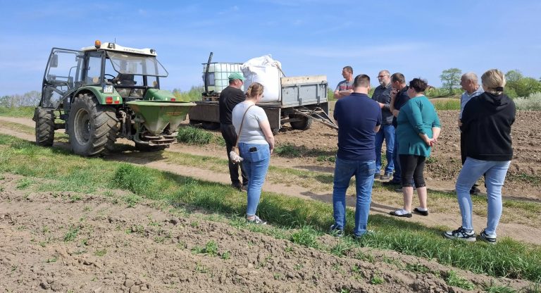 Grupa rolników na pokazie polowym w polu, słuchających prezentacji o mikroorganizmach w glebie.