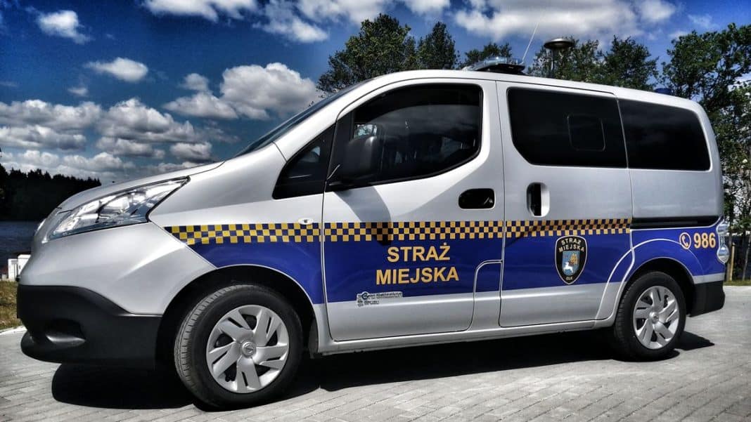 Samoch&oacute;d straży miejskiej zaparkowany na placu przy szkole, w tle chmury na niebie.