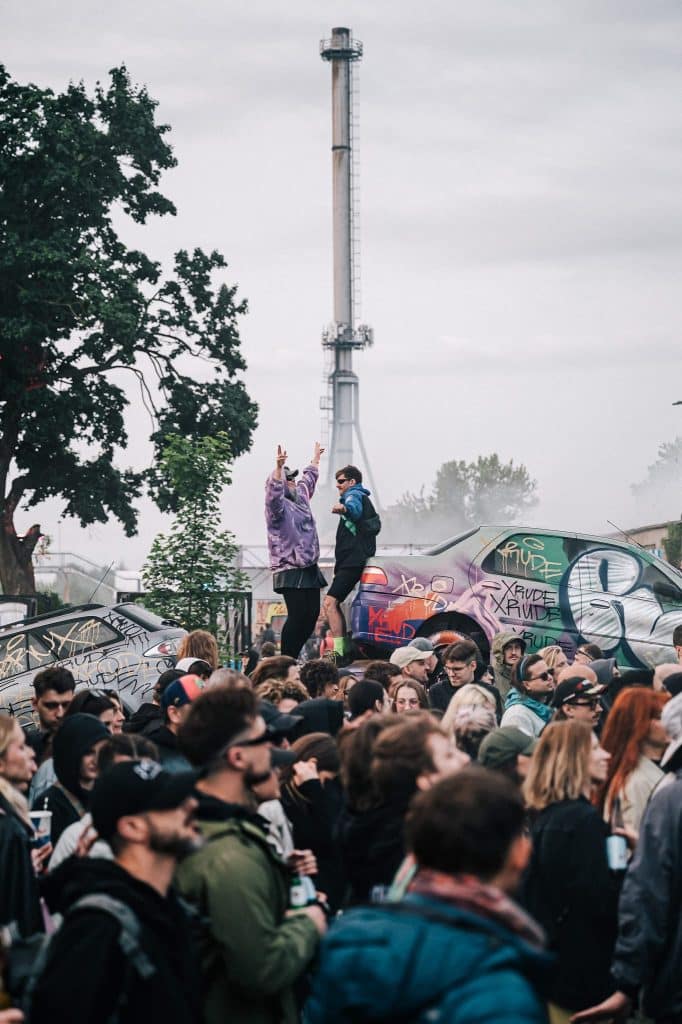 Tłum ludzi na festiwalu z graffiti na samochodach i przemysłową wieżą w tle.