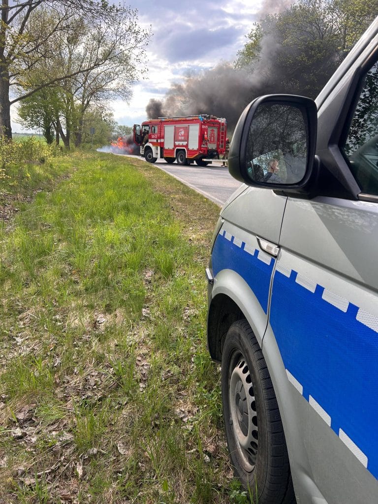 Samoch&oacute;d osobowy płonie na drodze, w tle straż pożarna gasząca pożar, z widokiem na niebo i drzewa.