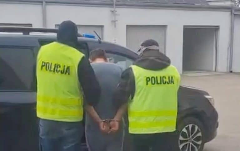 Mężczyzna w rękach policji, z tyłu radiowóz, na tle szarego budynku.