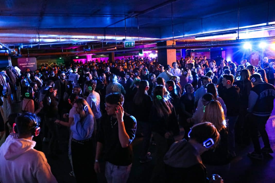 Tłum ludzi na parkingu Galerii Warmińskiej podczas Silent Party, z słuchawkami na głowach, podświetlonymi światłami.