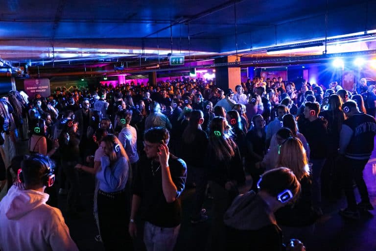 Tłum ludzi na parkingu Galerii Warmińskiej podczas Silent Party, z słuchawkami na głowach, podświetlonymi światłami.
