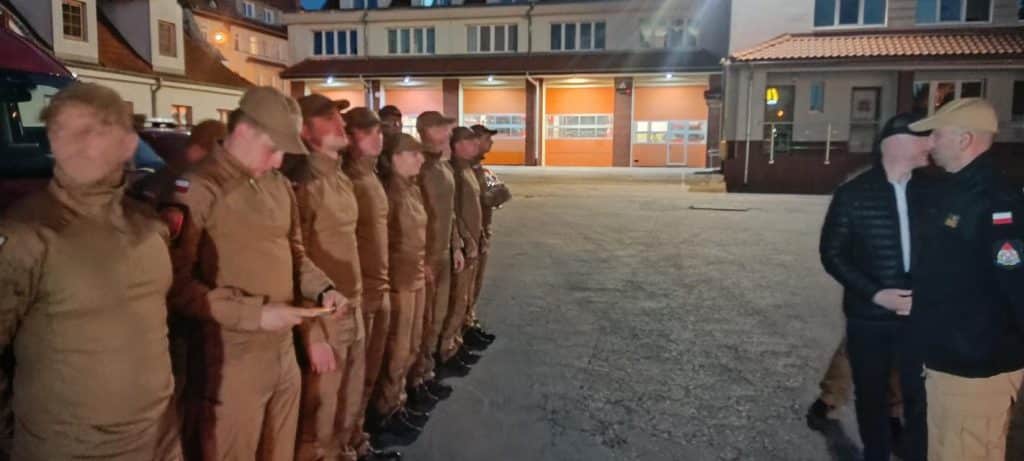 Grupa strażaków w uniformach stojąca w rzędzie na placu, rozmawiająca z innym funkcjonariuszem.