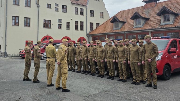 Grupa strażak&oacute;w z wojew&oacute;dztwa warmińsko-mazurskiego w uniformach stojąca przed pojazdami ratowniczymi na ulicy.