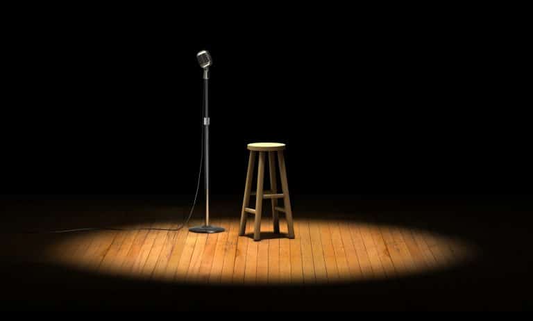 Scena stand-up z mikrofonem i stołkiem na tle ciemnej sceny, oświetlonej pojedynczym światłem.