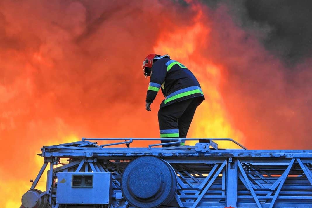 Strażak na dachu budynku podczas gaszenia pożaru z dużymi płomieniami w tle.