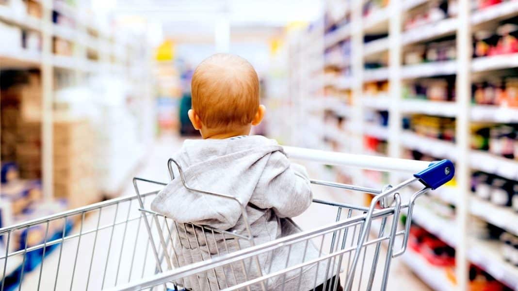 Małe dziecko siedzi w w&oacute;zku sklepowym, patrząc na p&oacute;łki pełne produkt&oacute;w spożywczych.