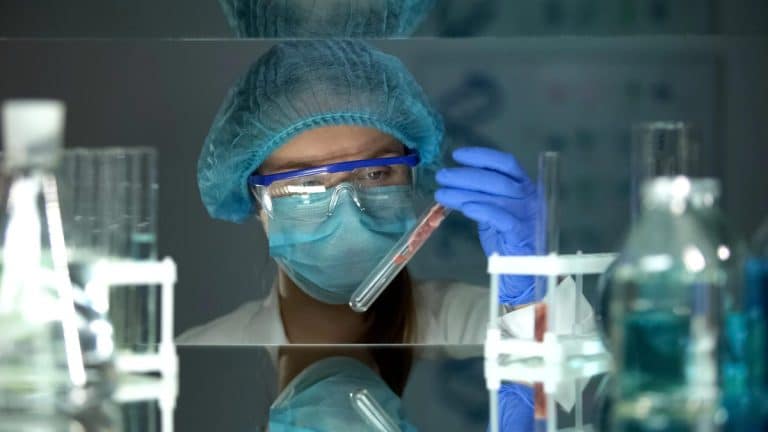 Laborantka w laboratorium bada próbkę, ubrana w maskę, okulary i rękawice, otoczona sprzętem laboratoryjnym.